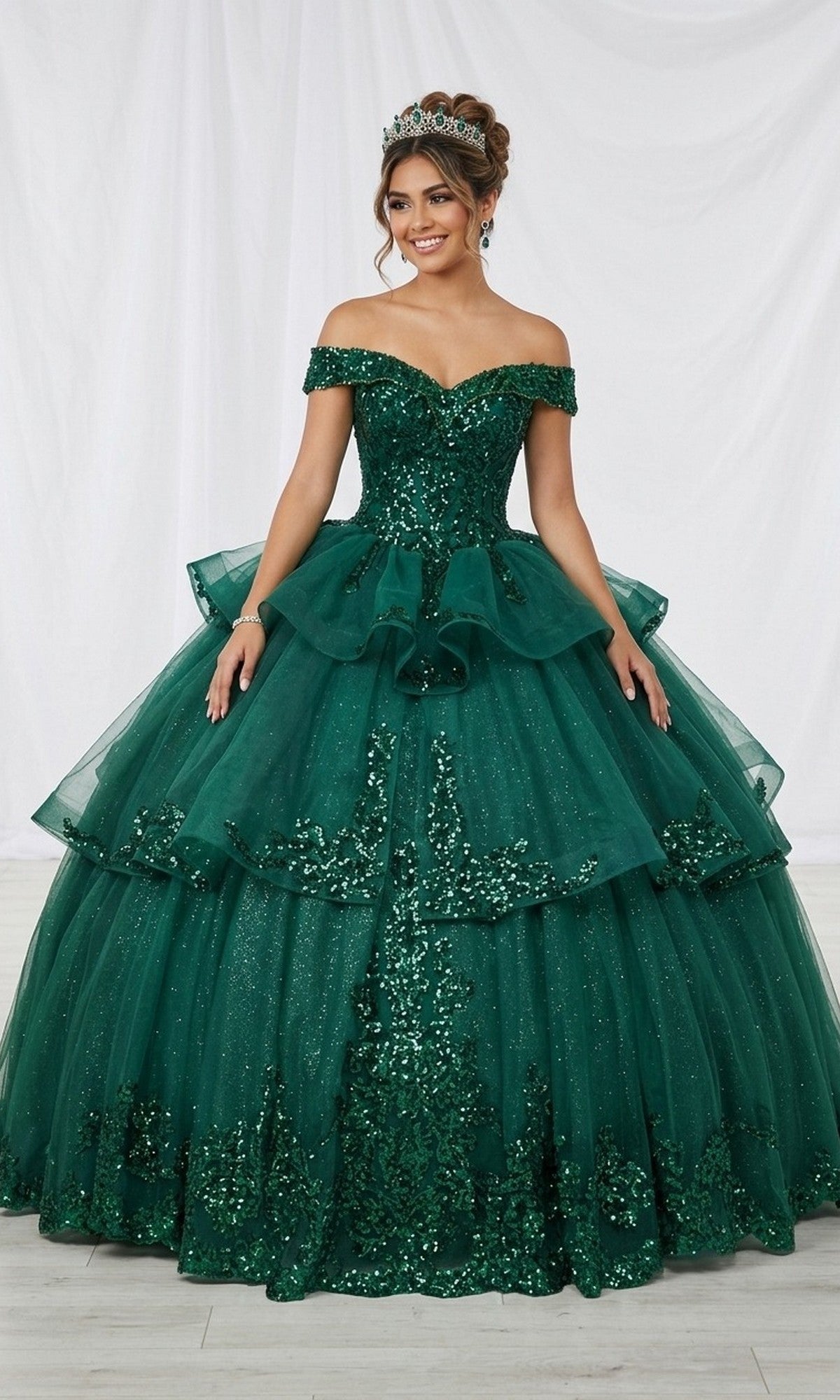 A woman wearing the Quinceanera Dress CHF3063 By Chicas in green, featuring a tiered ball gown silhouette with an off-shoulder neckline and sparkling sequin and tulle embellishments, stands against a white fabric backdrop.